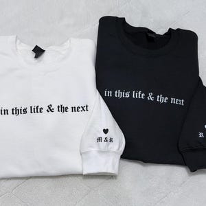 May include: Two sweatshirts, one white and one black, are displayed. Both have the phrase "in this life & the next" embroidered in white. The white sweatshirt has "M&R" and a heart on the cuff. The black sweatshirt has "R&M" and a heart on the cuff.