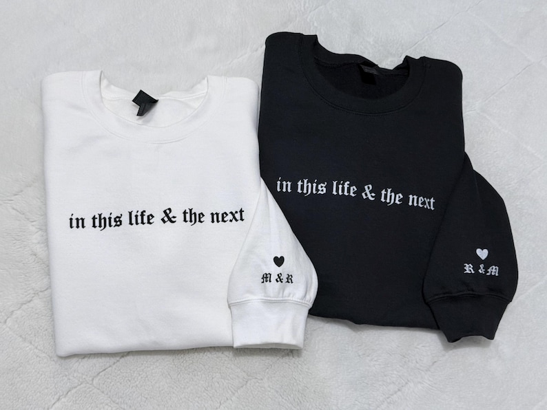 May include: Two sweatshirts, one white and one black, are displayed. Both have the phrase "in this life & the next" embroidered in white. The white sweatshirt has "M&R" and a heart on the cuff. The black sweatshirt has "R&M" and a heart on the cuff.