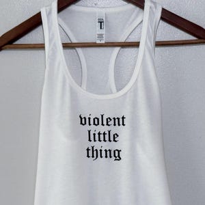 May include: White racerback tank top with the phrase "violent little thing" printed in black gothic font. The tank top is hanging on a wooden hanger against a neutral background. The shirt has a scoop neck and a racerback design.