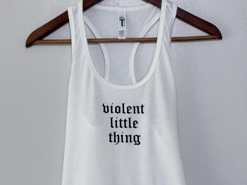 May include: White racerback tank top with the phrase "violent little thing" printed in black gothic font. The tank top is hanging on a wooden hanger against a neutral background. The shirt has a scoop neck and a racerback design.