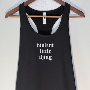 May include: Black racerback tank top with the words "violent little thing" in white gothic font. The tank top is hanging on a wooden hanger against a neutral background. The top has a scoop neck and a racerback design.