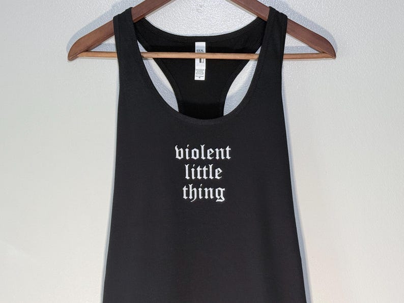 May include: Black racerback tank top with the words "violent little thing" in white gothic font. The tank top is hanging on a wooden hanger against a neutral background. The top has a scoop neck and a racerback design.