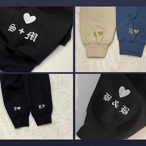 May include: Close-up of four different sweatshirt sleeves. Each sleeve features embroidered initials and a heart symbol. Colors include black, tan, and blue. The initials are in a gothic font.