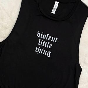 May include: Black sleeveless shirt with the text "violent little thing" in white gothic font. The shirt is made of a soft material and is likely for casual wear.