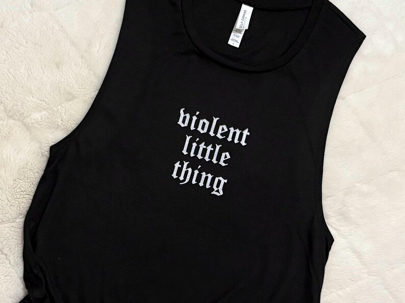 May include: Black sleeveless shirt with the text "violent little thing" in white gothic font. The shirt is made of a soft material and is likely for casual wear.