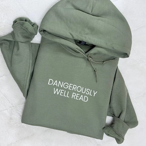 May include: An olive green hooded sweatshirt featuring long sleeves and a drawstring hood. The front of the garment displays the text DANGEROUSLY WELL READ printed in white capital letters across the center of the chest area.