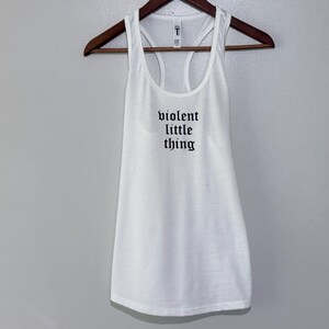 May include: White racerback tank top with the text "violent little thing" in black gothic font. The tank top is hanging on a wooden hanger against a light gray background. The tank top has a scoop neck and a racerback design.
