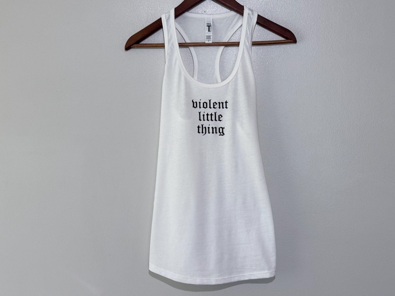 May include: White racerback tank top with the text "violent little thing" in black gothic font. The tank top is hanging on a wooden hanger against a light gray background. The tank top has a scoop neck and a racerback design.