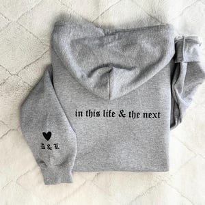 May include: A gray hoodie with a hood, featuring the text "in this life & the next" on the back in a gothic font. The left sleeve has a black heart and the initials "B & L". The hoodie is made of a soft material.