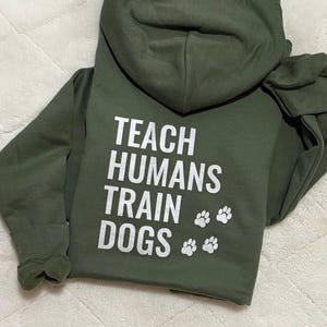 May include: An olive green hoodie with the white text "TEACH HUMANS TRAIN DOGS" and paw prints. The hoodie features long sleeves and a hood.