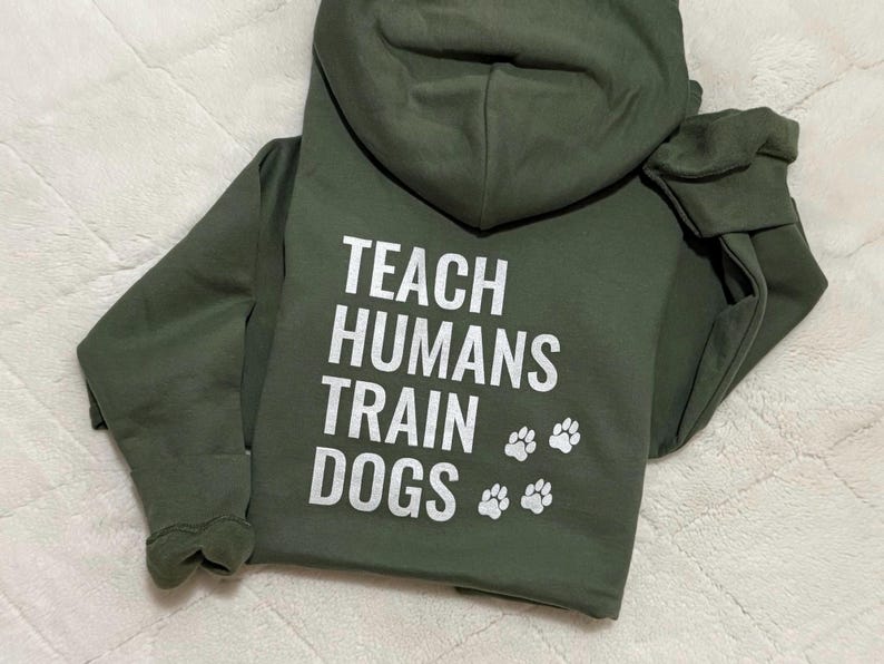 May include: An olive green hoodie with the white text "TEACH HUMANS TRAIN DOGS" and paw prints. The hoodie features long sleeves and a hood.