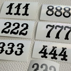 May include: A collection of white rectangular stickers with black numbers printed on them. The numbers include 111, 222, 333, 444, 777, and 888. The numbers are in a bold, decorative font.