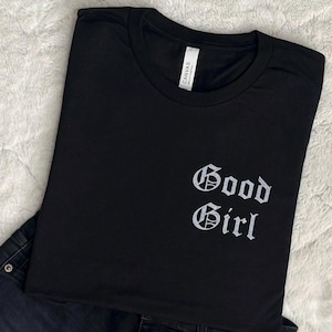 May include: Black t-shirt with the words "Good Girl" in a gothic font. The shirt is folded, with a tag visible at the neckline. Dark denim jeans are partially visible in the image.