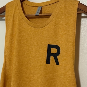 May include: Mustard yellow tank top with a black letter "R" on the front. The sleeveless shirt is hanging on a wooden hanger. The fabric has a heathered texture.