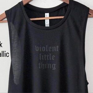 May include: Black sleeveless shirt with the words "violent little thing" printed in metallic ink. The shirt is hanging on a wooden hanger against a neutral background. The text is in a gothic font.