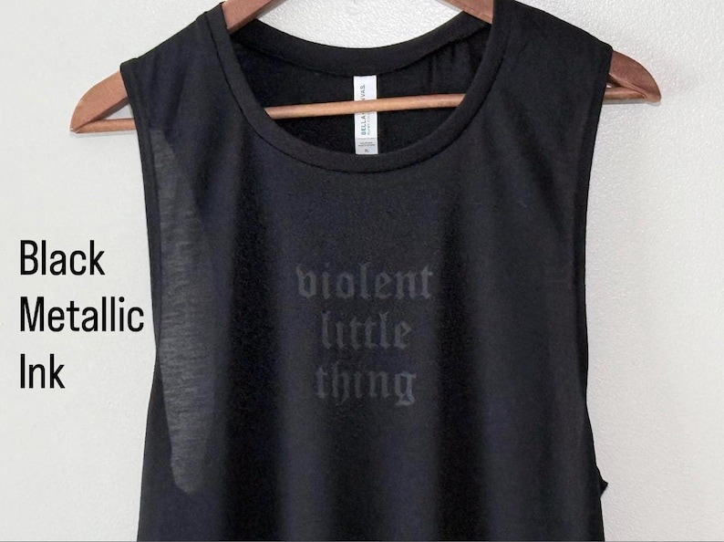 May include: Black sleeveless shirt with the words "violent little thing" printed in metallic ink. The shirt is hanging on a wooden hanger against a neutral background. The text is in a gothic font.