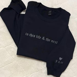 May include: Black long-sleeve sweatshirt with the embroidered text "in this life & the next" across the chest. The sleeve features a heart and the initials "J&M". The sweatshirt is folded on a white, textured surface.