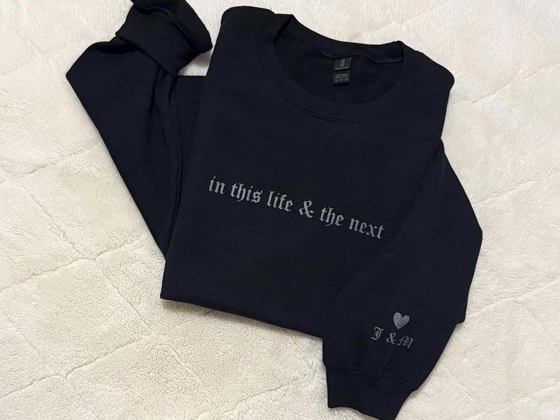 May include: Black long-sleeve sweatshirt with the embroidered text "in this life & the next" across the chest. The sleeve features a heart and the initials "J&M". The sweatshirt is folded on a white, textured surface.