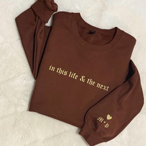 May include: A brown long-sleeve sweatshirt with the embroidered text "in this life & the next" in a gold color. The sleeve features gold initials and a heart. The sweatshirt is folded on a white, textured surface.