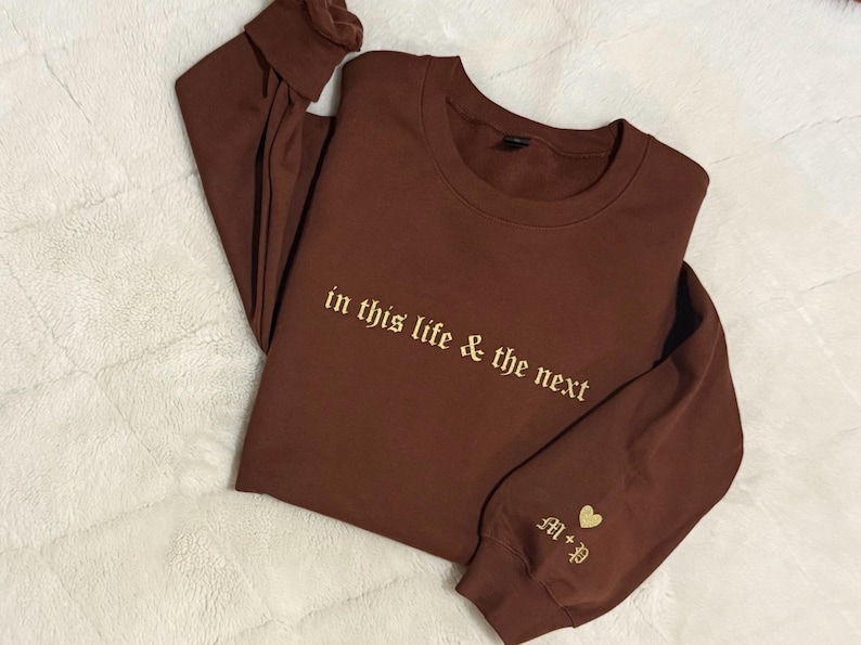 May include: A brown long-sleeve sweatshirt with the embroidered text "in this life & the next" in a gold color. The sleeve features gold initials and a heart. The sweatshirt is folded on a white, textured surface.