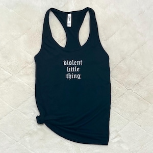 May include: Black racerback tank top with the phrase "violent little thing" in white gothic font. The tank top is made of a soft material and is knotted at the bottom. The background is a textured, off-white surface.
