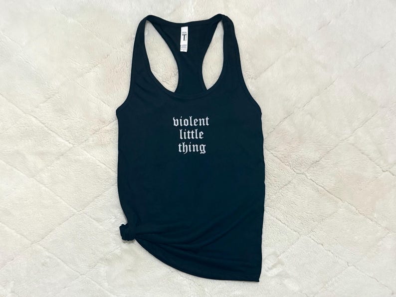 May include: Black racerback tank top with the phrase "violent little thing" in white gothic font. The tank top is made of a soft material and is knotted at the bottom. The background is a textured, off-white surface.