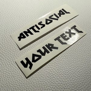 May include: Two white rectangular stickers with black text. The top sticker reads "ANTISOCIAL" and the bottom sticker reads "YOUR TEXT". The stickers are on a textured, off-white surface.