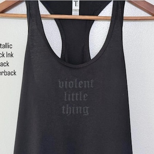 May include: Black racerback tank top with the words "violent little thing" printed on the front in metallic black ink. The tank top is hanging on a wooden hanger. The text "Metallic Black Ink Black Racerback" is printed on the left side.