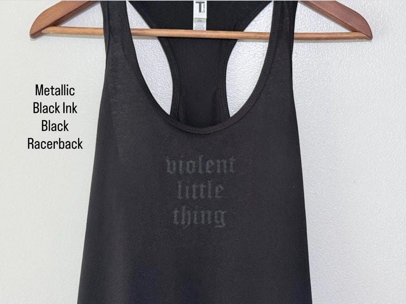May include: Black racerback tank top with the words "violent little thing" printed on the front in metallic black ink. The tank top is hanging on a wooden hanger. The text "Metallic Black Ink Black Racerback" is printed on the left side.