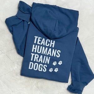 May include: A navy blue hoodie featuring the white text "TEACH HUMANS TRAIN DOGS" and paw prints. The hoodie is folded, displaying the back design. A casual, comfortable garment.
