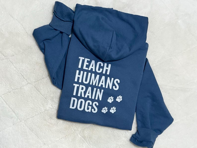 May include: A navy blue hoodie featuring the white text "TEACH HUMANS TRAIN DOGS" and paw prints. The hoodie is folded, displaying the back design. A casual, comfortable garment.