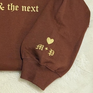 May include: A maroon sweatshirt sleeve with gold lettering that reads "life & the next". The sleeve cuff features a gold heart and the initials "M + P" in an elegant font. The fabric has a soft, textured appearance.