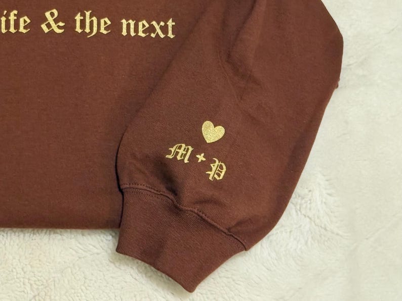 May include: A maroon sweatshirt sleeve with gold lettering that reads "life & the next". The sleeve cuff features a gold heart and the initials "M + P" in an elegant font. The fabric has a soft, textured appearance.