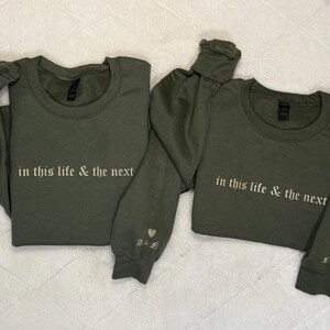 May include: Two olive green crewneck sweatshirts. The text "in this life & the next" is embroidered in white across the chest. One sleeve has a heart and initials embroidered. A matching set of apparel.