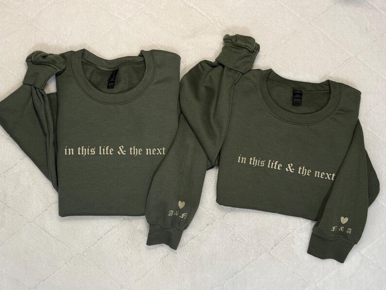May include: Two olive green crewneck sweatshirts. The text "in this life & the next" is embroidered in white across the chest. One sleeve has a heart and initials embroidered. A matching set of apparel.