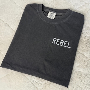 May include: Dark gray t-shirt with the word "REBEL" printed in white on the front. The shirt is made of a soft material and has a classic crew neck design. The shirt is folded neatly on a white, textured surface.