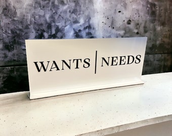 May include: White sign with black text that reads "WANTS | NEEDS".