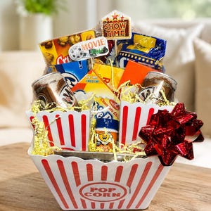 May include: A movie night gift basket in a red and white striped popcorn container filled with various snacks, two cans of A&W root beer, yellow paper shred, and a red bow. A sign at the top reads SHOW TIME and another reads MOVIE NIGHT.