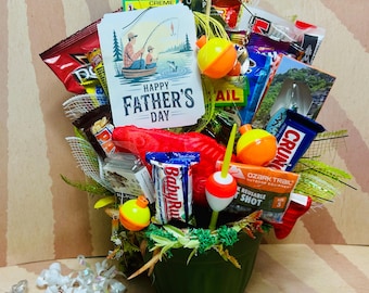Puede incluir: Una maceta verde rebosante de regalos temáticos de pesca, que incluyen caramelos, flotadores de pesca y una tarjeta que dice "Happy Father's Day". El arreglo está decorado con detalles verdes y amarillos y cintas de colores.