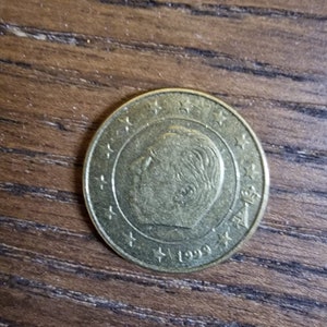 May include: A gold-colored coin with a portrait of a man on the front. The coin has the year 1999 and a crown on the back.