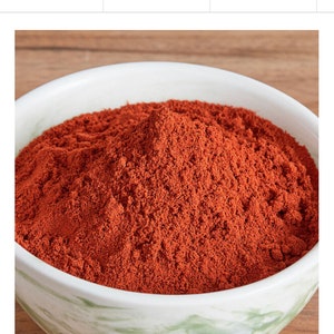 May include: A white and green marbled ceramic bowl filled with a mound of fine, reddish-brown powder. The powder has a textured appearance, and the bowl is set against a blurred wooden background.
