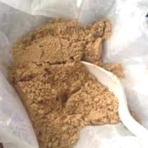 Può includere: Primo piano di una sostanza granulare marrone chiaro, possibilmente un prodotto alimentare, all'interno di un sacchetto di plastica trasparente. Un cucchiaio di plastica bianco è parzialmente visibile, suggerendo che la sostanza viene prelevata.