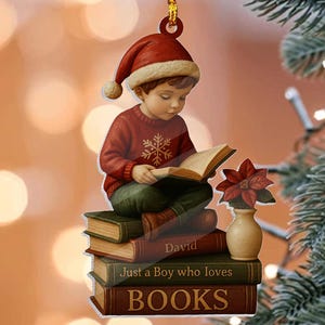 以下が含まれることがあります： サンタ帽をかぶった男の子が本を読んでいるクリスマスのオーナメント。本は積み重ねられており、「David」、「Just a Boy who loves」、「BOOKS」と書かれています。赤い花が入った小さな花瓶が本の隣にあります。