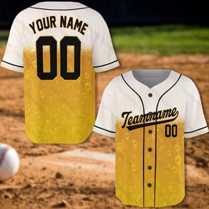 Pode incluir: Duas camisas de beisebol brancas com um design temático de cerveja. A frente das camisas apresenta um padrão de cerveja amarelo e dourado com bolhas. As camisas têm números e texto pretos. O texto na primeira camisa diz "YOUR NAME" e o texto na segunda camisa diz "Teamoname".