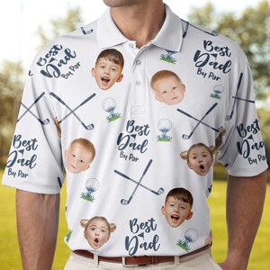 May include: White polo shirt with a repeating pattern of golf clubs, golf balls, and children's faces. The shirt also features the text "Best Dad By Par" in a navy blue script. The shirt has a collared neckline and short sleeves.