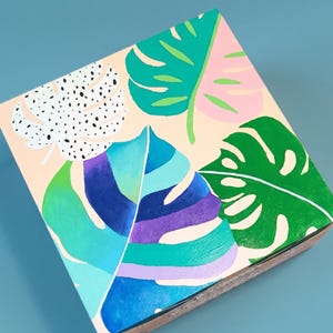 May include: A square wooden box featuring a hand-painted design of various tropical leaves on a light peach background. The artwork includes a white leaf with black dots, a solid green leaf, a pink leaf, and a large central leaf in blue and purple.