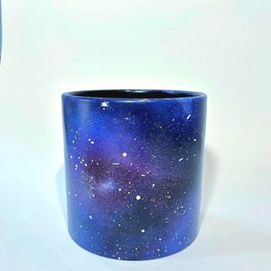 May include: A cylindrical ceramic pot with a galaxy design. The pot features a dark blue base that fades into a deep purple center, speckled with white dots. The interior is black.