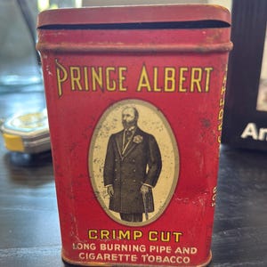 Puede incluir: Lata de tabaco Prince Albert vintage. La lata roja presenta la inscripción "Prince Albert" en letras doradas y un retrato ovalado de un hombre. La parte inferior de la lata dice "Crimp Cut Long Burning Pipe and Cigarette Tobacco."