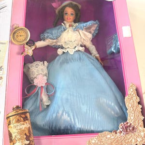 Puede incluir: Muñeca vintage en una caja rosa, vestida con un vestido azul claro con cuello de encaje blanco y sombrero a juego. La muñeca sostiene un pequeño ramo y un reloj de bolsillo dorado. La caja también contiene una lata de polvos de talco vintage.