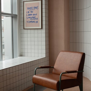 Pu&ograve; includere: Una stampa incorniciata con il testo "HOUSE RULES" e le frasi "BE YOU", "SHARE IDEAS", "OK to make MISTAKES" e "EMOTIONS OK". Una poltrona in pelle marrone con struttura in metallo verde &egrave; di fronte a un muro piastrellato.